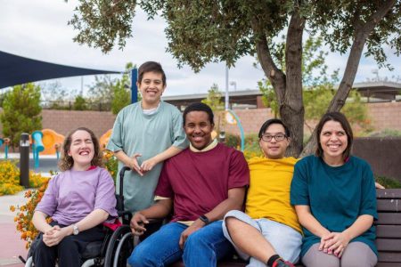 A diverse group of five adults sitting together outdoors on a bench and in a wheelchair, smiling at the camera under a tree with playground equipment visible in the background.