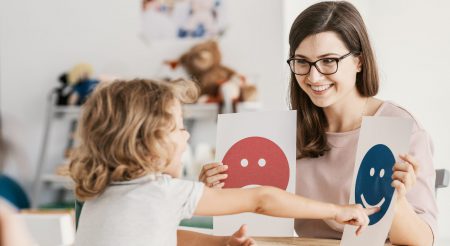 Young child with curly hair points at a smiling blue face on a card while a smiling adult woman wearing glasses holds two cards, one with a sad red face and one with a happy blue face, in a bright room with toys and stuffed animals in the background