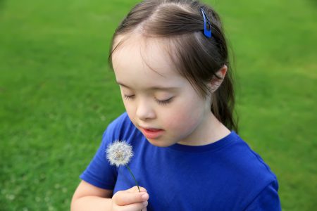 Young girl with Down syndrome wearing a blue shirt holds and looks at a dandelion seed head against a blurred green grassy background.