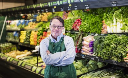 Smiling grocery store worker wearing glasses and a green apron stands with arms crossed in front of a colorful produce display of lettuce, peppers, and other vegetables.