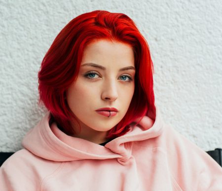Close-up portrait of a teenage girl with bright red hair and light blue eyes, looking directly at the camera with a neutral expression. She is wearing a light pink hooded sweatshirt and has a small lip piercing on the lower lip. The background is a light textured white wall, and the focus is on her face and upper shoulders.