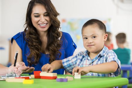 A smiling woman with long brown hair sits at a table with a young boy who has Down syndrome; they play with colorful wooden blocks together in a bright, classroom-like setting.