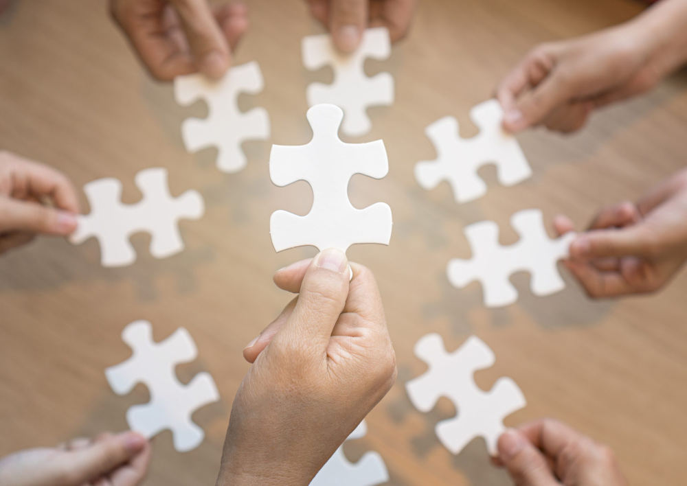 Eight hands hold big, white puzzle pieces. One hand holds a puzzle piece in the center foreground. In the slightly blurred background, four people hold puzzle pieces above a tan, brown table. You can't see one of the hands, which is below the foreground hand. But you can see 8 puzzle pieces in the background. The total number of puzzle pieces is nine.